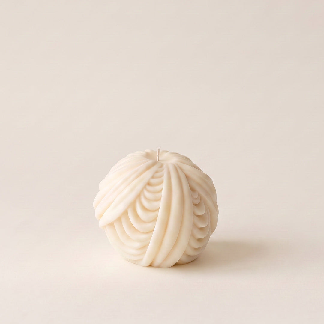 An off-white, textured spherical candle with a ribbed, pleated pattern, set against a solid, warm off-white background.