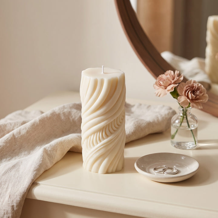A tall cream-colored pillar candle with a sculpted, wavy ribbed texture stands on a light beige table. The scene is styled with a draped linen cloth, a small glass vase with pink flowers, a ceramic ring dish, and a wooden mirror in the background.