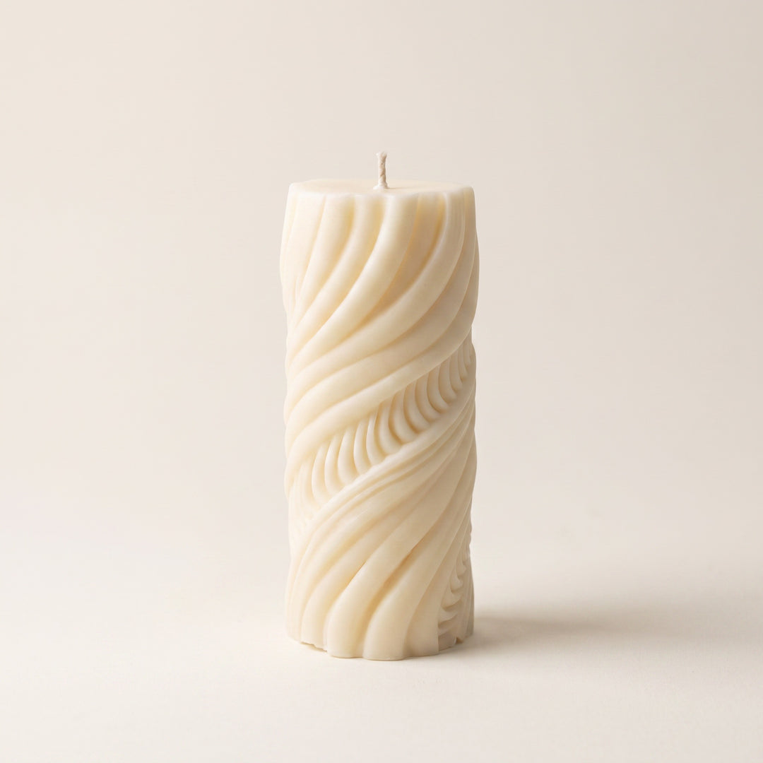 An off-white pillar candle with a sculpted, twisted wave pattern, standing upright on a neutral cream background.