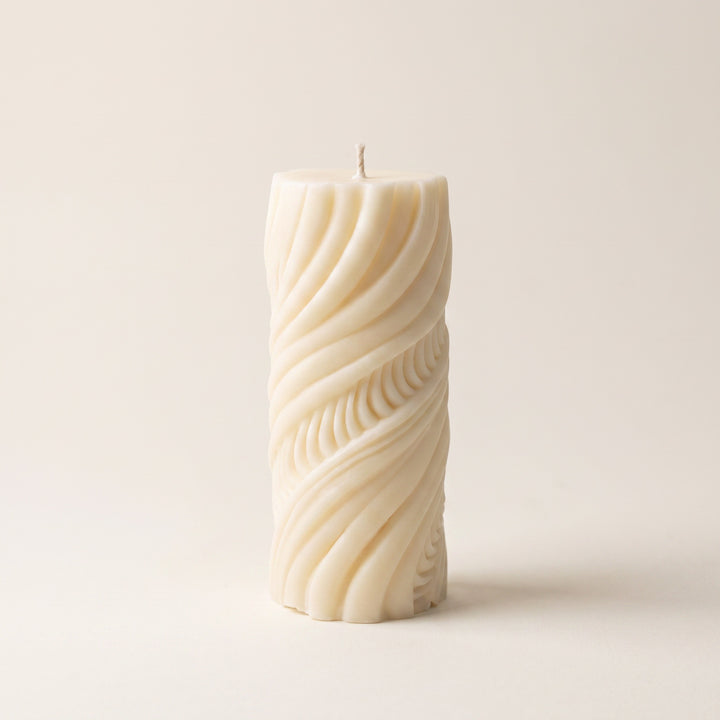 An off-white pillar candle with a sculpted, twisted wave pattern, standing upright on a neutral cream background.