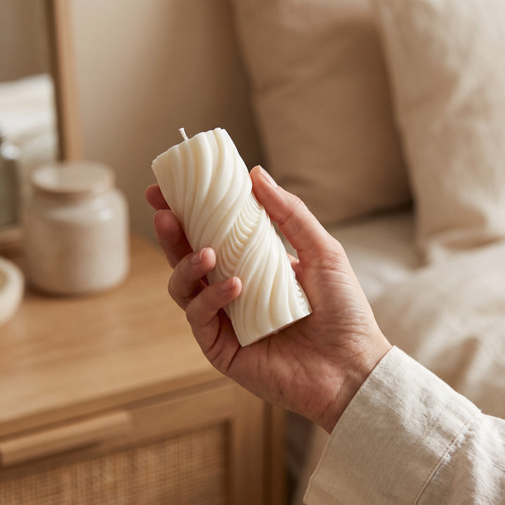 A hand holding a cream-colored twisted pillar candle in a cozy, warm-toned bedroom setting.