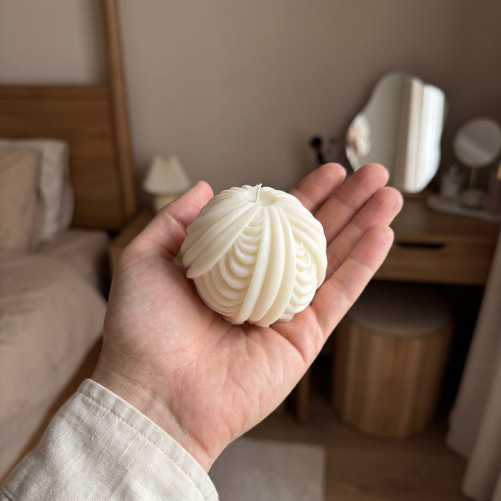 A hand holding a ribbed, pumpkin-shaped cream candle in a cozy bedroom setting.