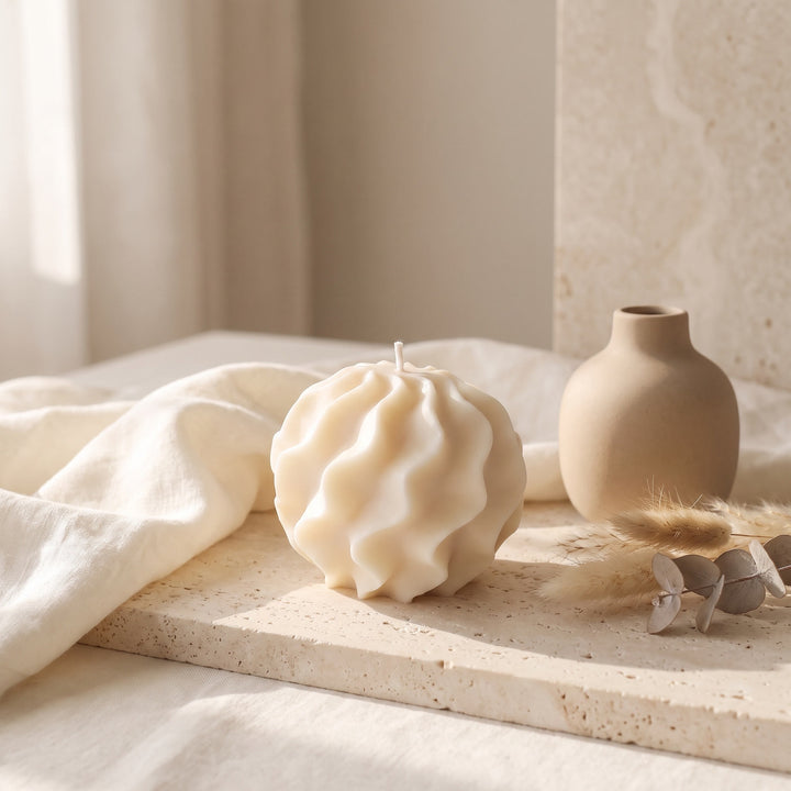 A sculptural, textured cream-colored candle sits on a stone tray next to a small beige ceramic vase and dried botanicals, set against a soft neutral background with draped fabric.