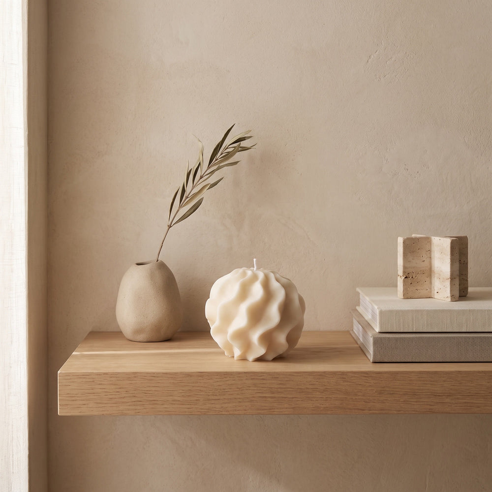A textured cream-colored sculptural candle sits on a light wood floating shelf, styled with a small beige ceramic vase holding a dried olive branch and a stack of neutral books.
