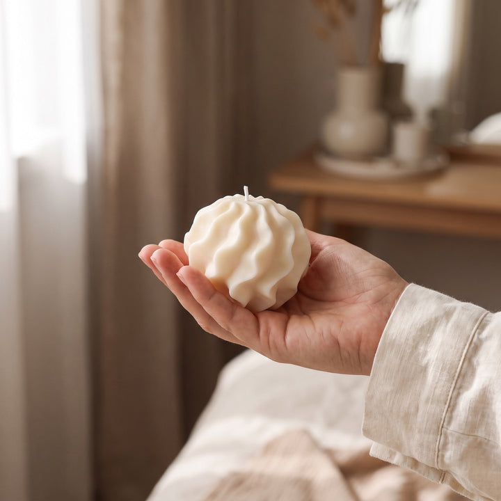 A hand holding a textured, pleated cream-colored candle in a softly lit, cozy indoor setting.