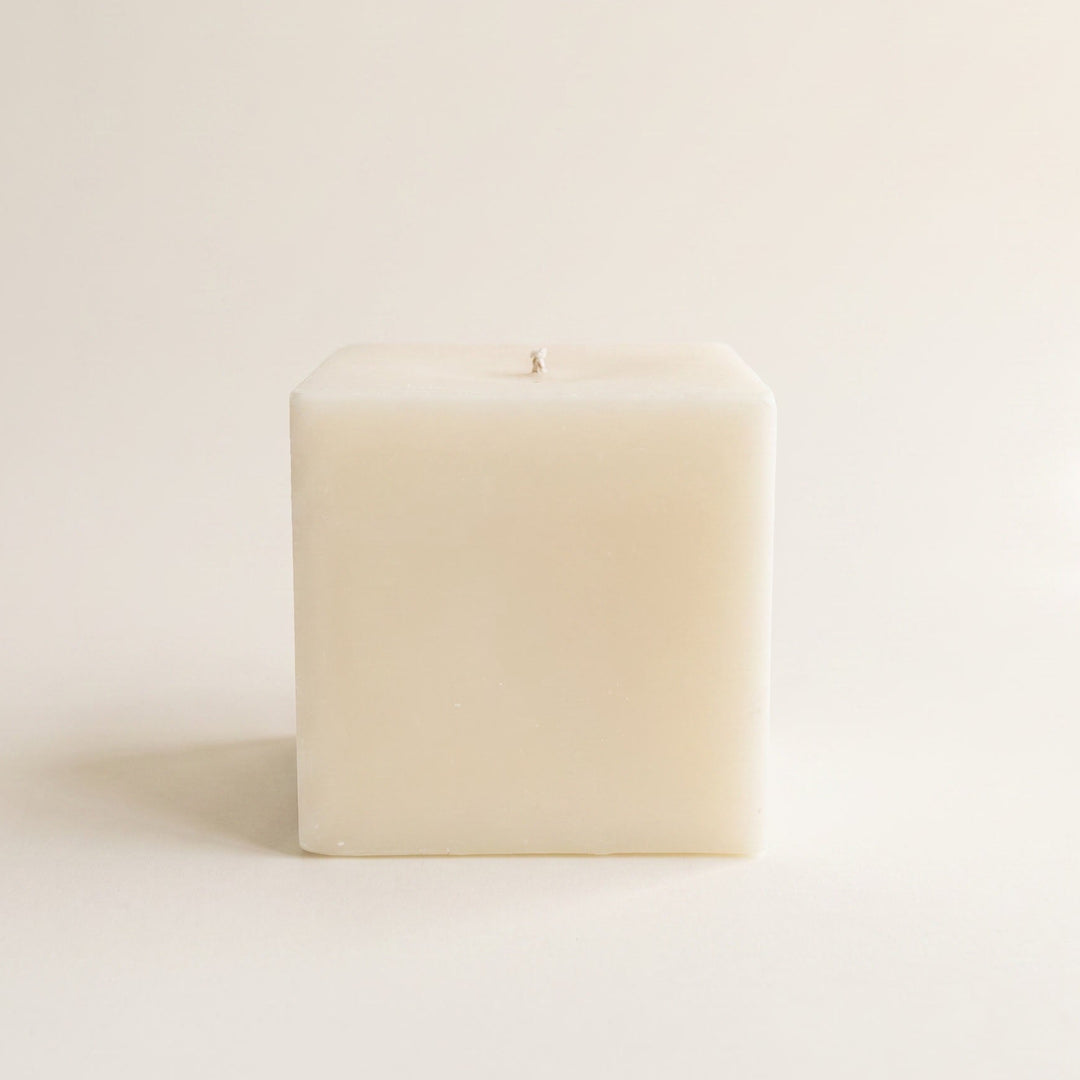 A single square cream-colored candle with a wick on top, photographed against a seamless, warm off-white background.