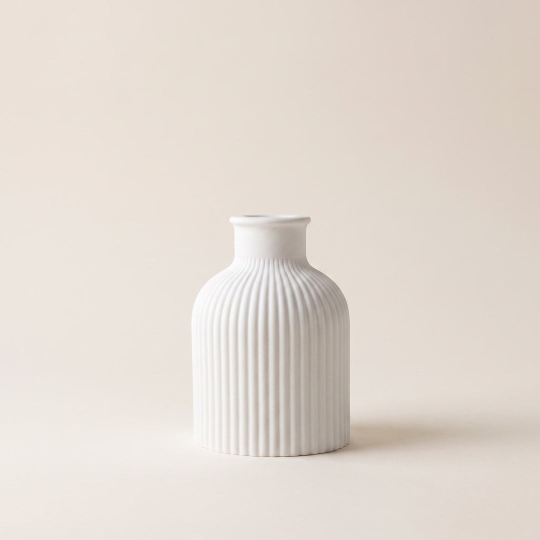 A small white ceramic vase with a fluted, vertically ribbed body and a short, narrow neck, centered against a solid, warm off-white background.