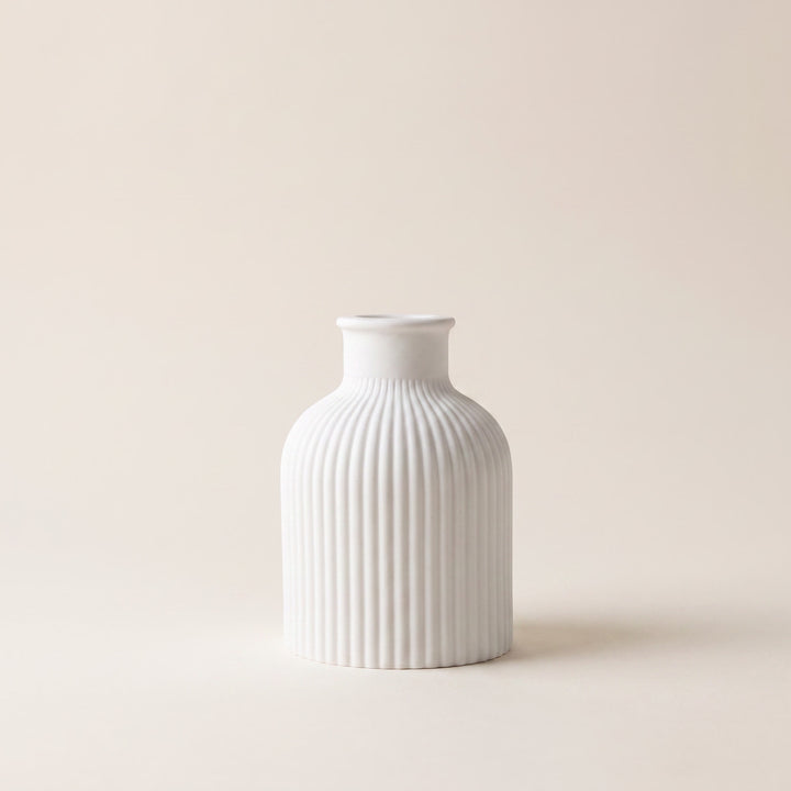 A small white ceramic vase with a fluted, vertically ribbed body and a short, narrow neck, centered against a solid, warm off-white background.