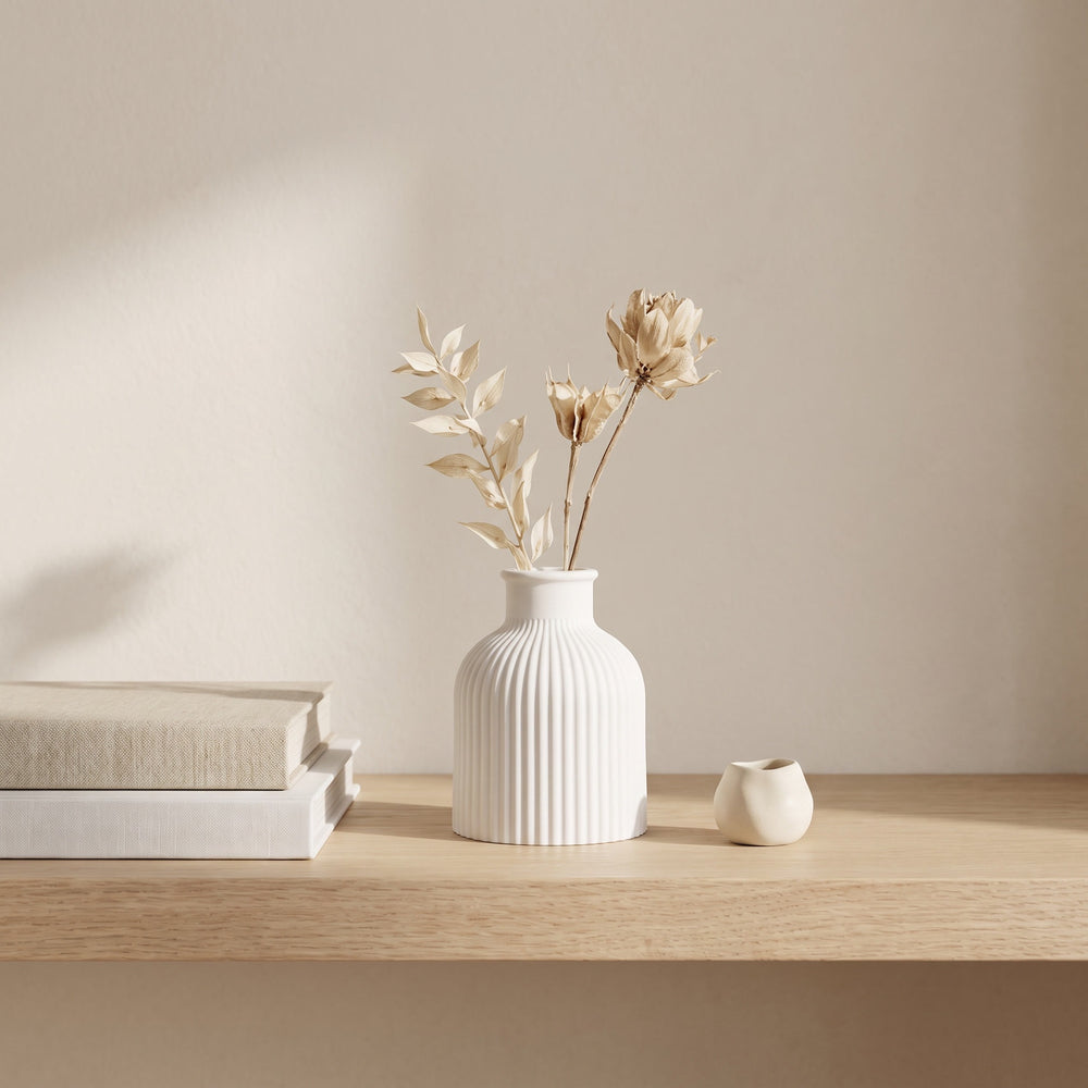 A white ribbed ceramic vase with dried beige flowers, styled on a light wood shelf next to a small white ceramic pot and stacked books against a warm beige wall.