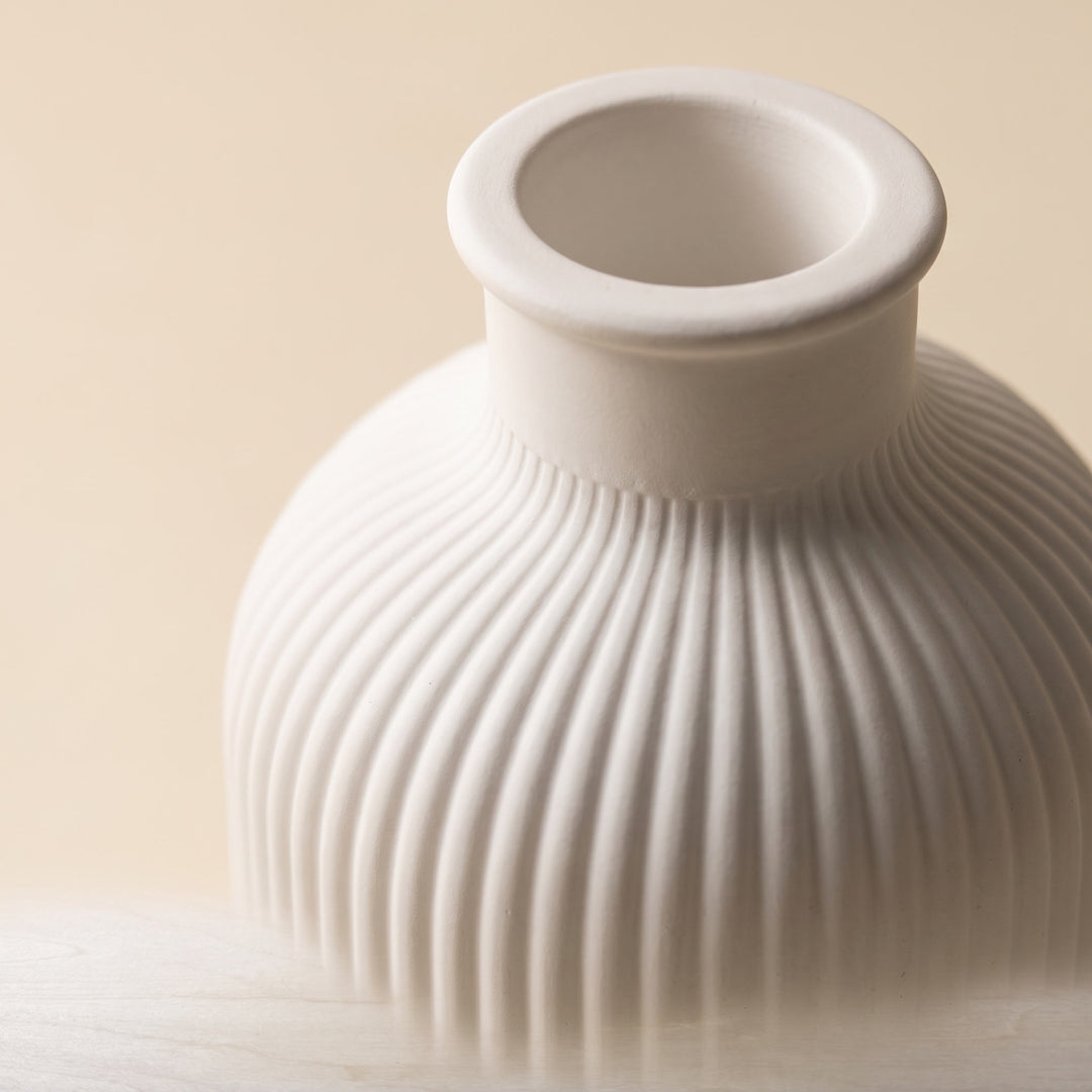 A close-up of a ribbed ceramic vase in a warm off-white or cream color, set against a solid beige background.