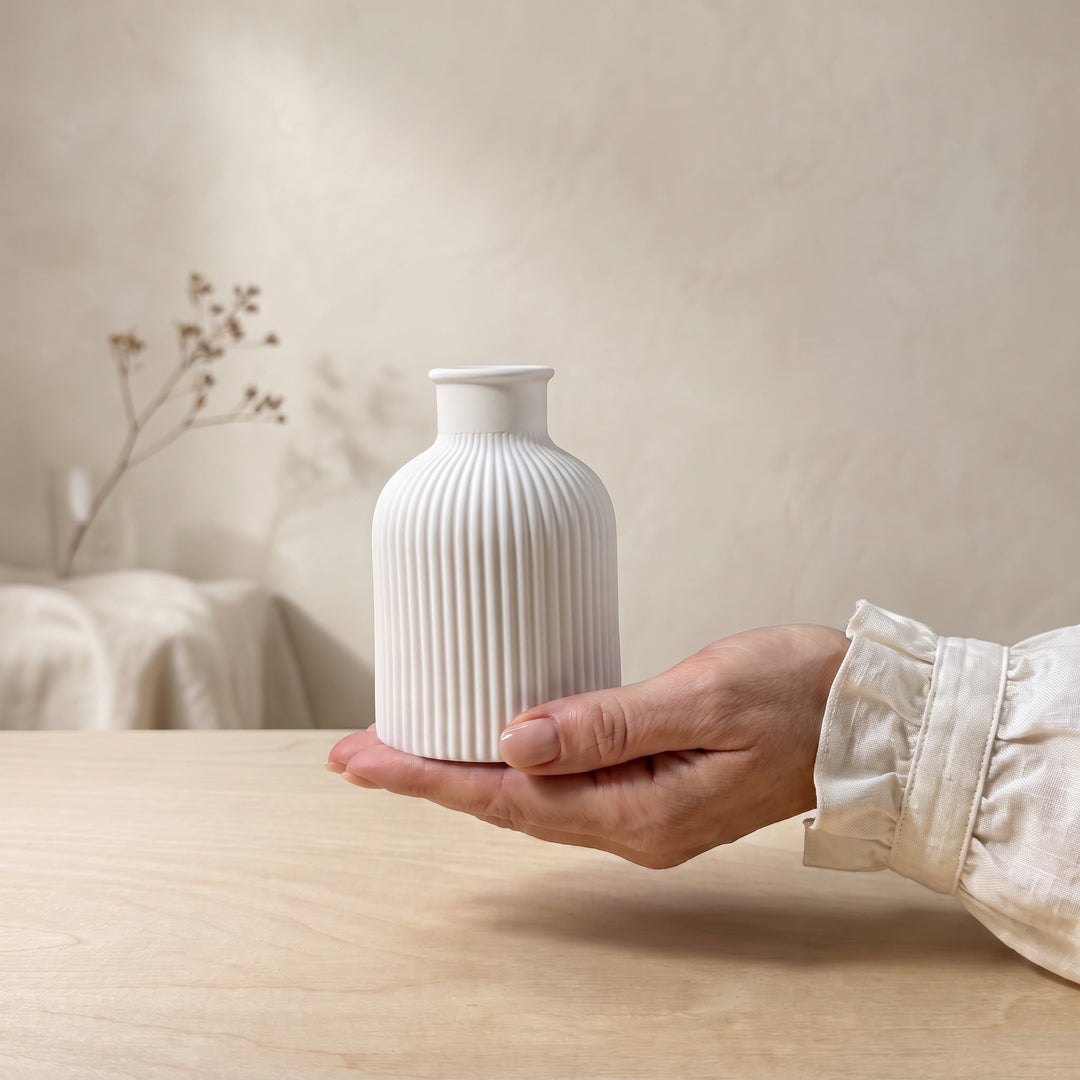 A hand holding a small white ribbed ceramic vase on a light wood table, with a softly blurred neutral interior background.