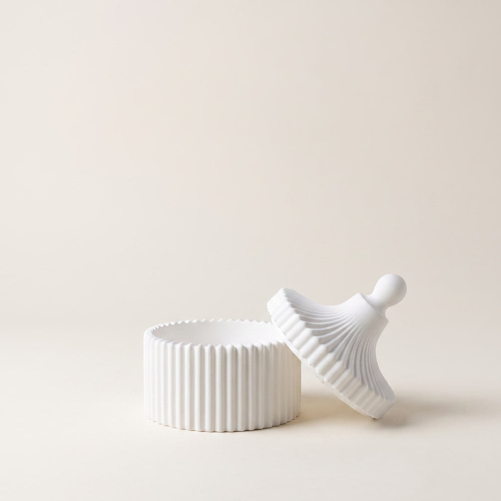 A white ribbed ceramic lidded container with its matching lid leaning against it, set against a solid warm beige background.