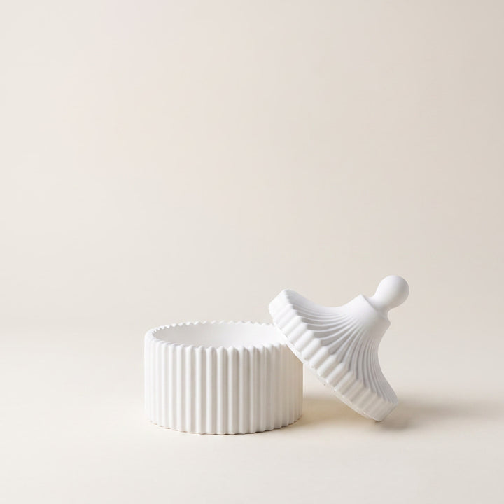 A white ribbed ceramic lidded container with its matching lid leaning against it, set against a solid warm beige background.