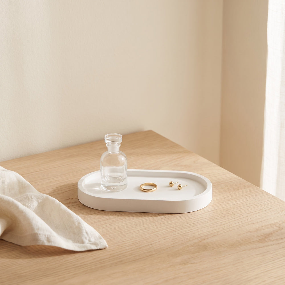 A white oval jewelry tray with a clear glass bottle and gold jewelry pieces, styled on a light wood table next to a cream linen cloth.