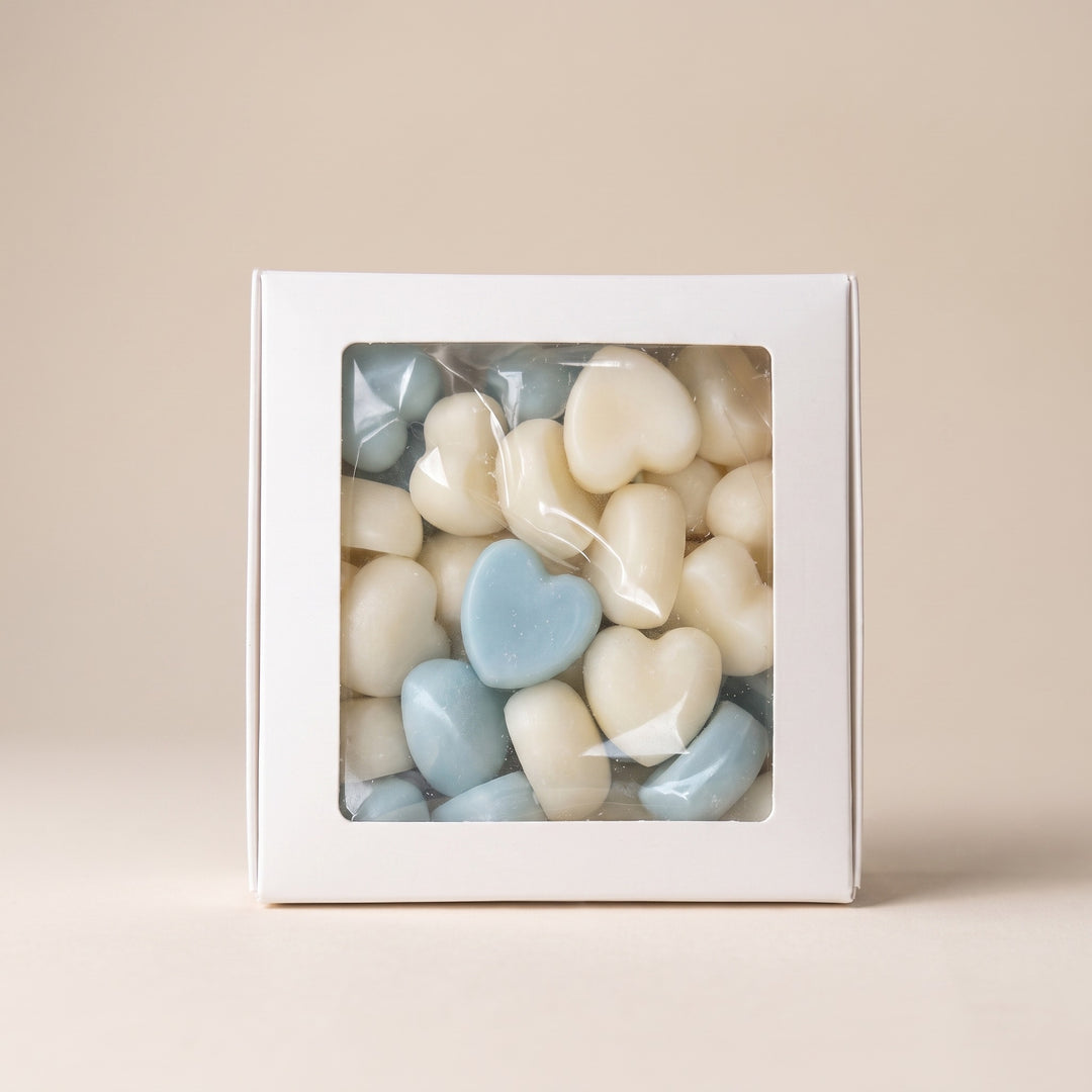 A white square gift box with a clear plastic window showing small heart-shaped wax melts in cream and light blue colors, set against a solid beige background.